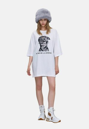 Jeune femme portant une grande robe t-shirt blanche avec un imprimé chien, un chapeau gris en fausse fourrure et des baskets blanches montantes avec des accents marron.