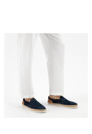 Person trägt weiße Hose und marineblaue Slipper mit braunen und beigen Sohlen-Details, steht auf einer weißen Oberfläche.