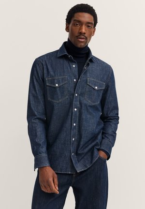 Denim shirt in dark blue with a classic collar, two chest pockets, and button closure. Features white stitching and a relaxed fit.