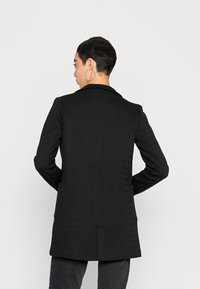 Blazer nero con una texture liscia, collo a rever e due tasche laterali. La parte posteriore presenta un dettaglio di cucitura verticale e uno spacco centrale.