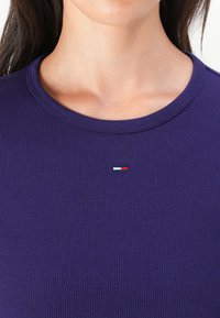 Violet juostos viršus turi apvalų iškirpimą ir mažą logotipą raudona, balta ir mėlyna spalvomis centre, demonstruojantį prigludusią dizainą ir glotnią tekstūrą.