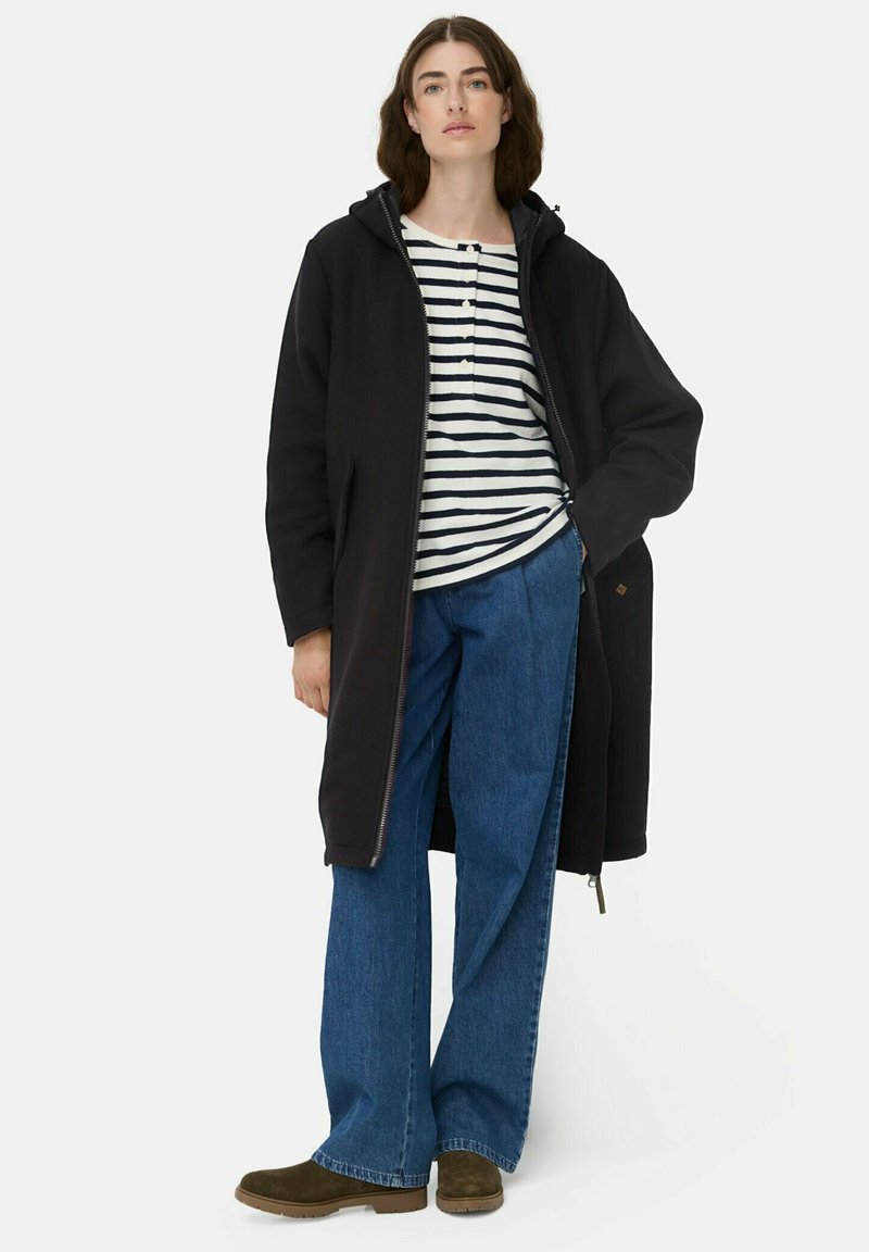 Schwarze Kapuzenjacke, gestreiftes Langarmshirt, blaue weit geschnittene Jeans und braune Wildleder-Stiefeletten. Neutrale Hintergrund.
