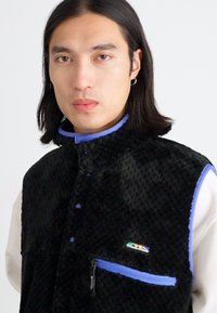 Black fleece vest with a textured surface, blue accents, high collar, snap buttons, and a zippered pocket on the front.