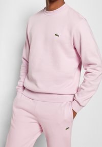 Felpa rosa in cotone con scollo rotondo e polsini a costine, con un piccolo logo di coccodrillo verde sul petto. Abbinata a pantaloni jogger coordinati.