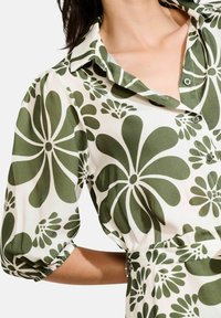 Camicia a bottoni floreale in crema e verde, con grandi motivi di fiori astratti. Il tessuto ha una texture liscia con maniche a palloncino.
