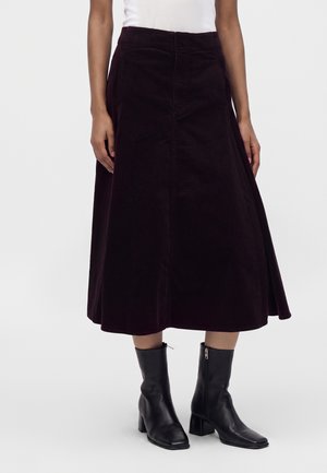 Femme portant une jupe midi violet foncé avec un top blanc et des bottines noires à talons, debout devant un fond clair uni.