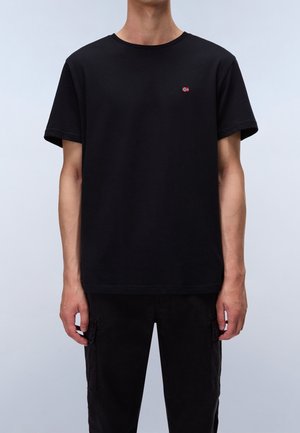 Homme portant un t-shirt noir uni à manches courtes avec un petit logo de drapeau rouge et blanc sur la poitrine gauche, associé à un pantalon noir.