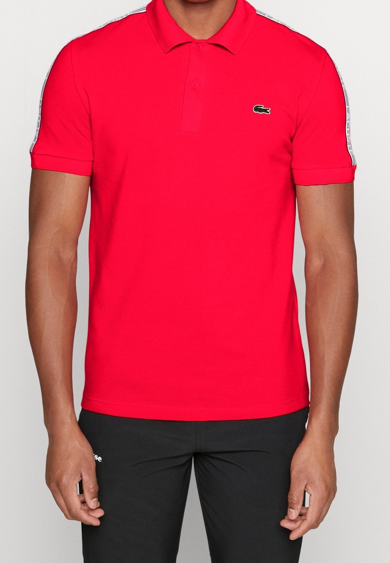 Homme portant un polo rouge vif avec un petit logo crocodile vert sur la poitrine et un pantalon noir sur un fond uni.