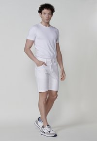 Bijela pamučna majica, bijele traperice, i sivi sportski tenisice sa plavim detaljima. Model stoji s jednom rukom u džepu, minimalna pozadina.