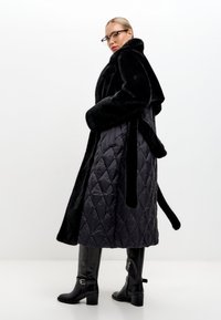 Zwarte gewatteerde lange jas met nepbont kraag en mouwen, met een ceintuur in de taille, gecombineerd met zwarte kniehoge haklaarzen.