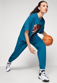 Sieviete teal sporta apģērbā driblo oranžo basketbola bumbu, ģērbusies baltās un melnās sporta kurpes, pozējusi gatava kustībai uz vienkrāsaina fona.