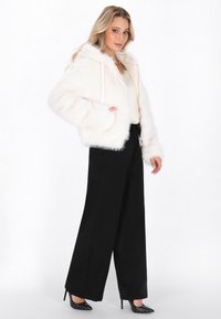 Faux-Fur-weiße Jacke mit Kapuze und Kordelzügen; lange schwarze Schlaghosen; schwarze spitze Absätze mit weißen Punkten; heller Hintergrund.