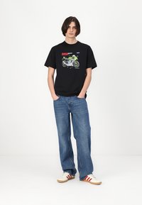 Zwarte t-shirt met korte mouwen met een motorfietsprint, gecombineerd met lichtblauwe losse jeans en crèmekleurige sneakers met rode accenten.