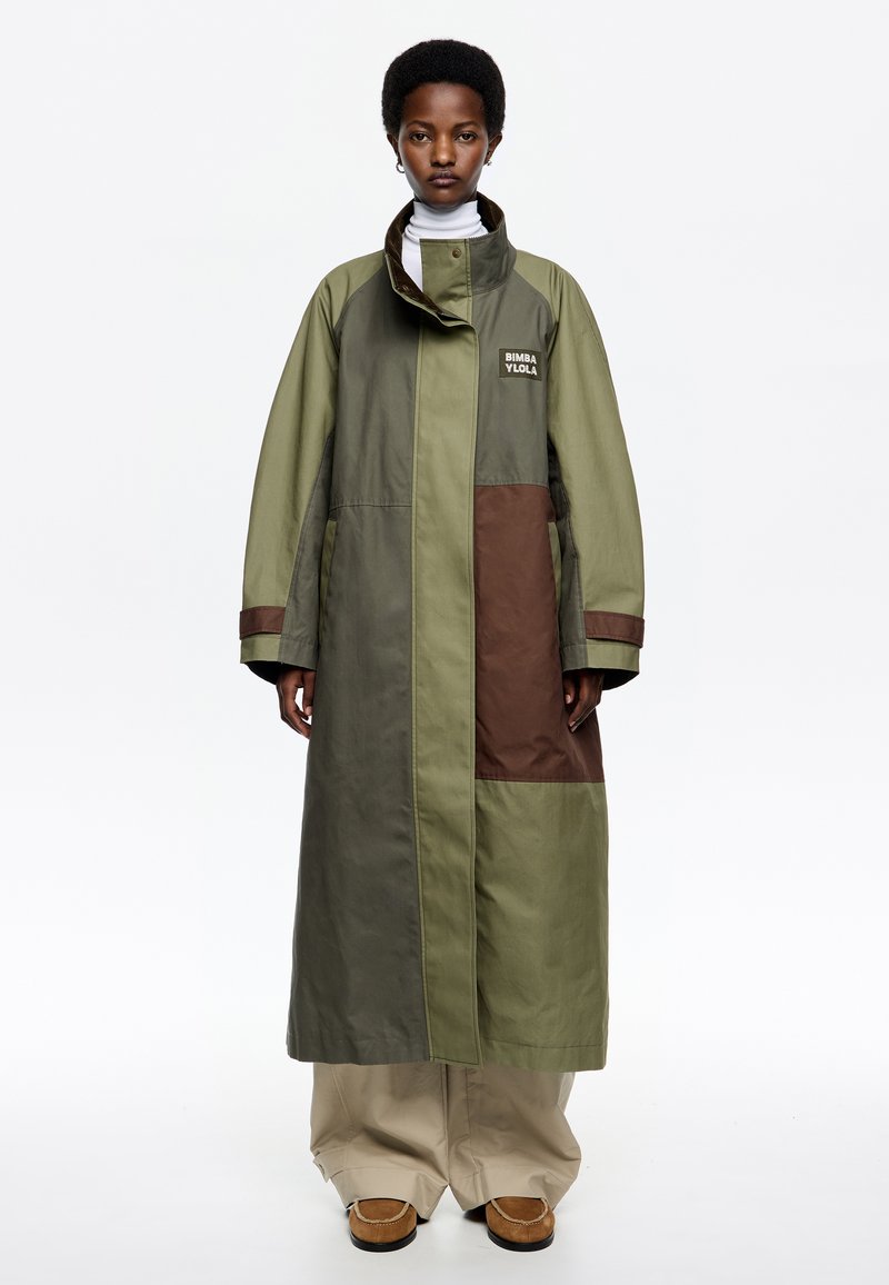Abrigo largo y oversize en tonos verde oliva y marrón, con un cuello alto, cierre de cremallera y bolsillos laterales. Logo en el pecho.
