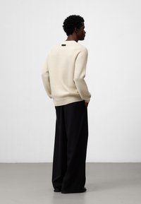 Beige stickad tröja med texturerad yta, rundhalsringning och svart märke på baksidan, kombinerad med vida svarta byxor.