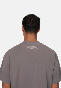 Grijze oversized t-shirt van zacht katoen. Heeft een ronde halslijn en witte bedrukte tekst op de rug, met de merknaam en het jaar van oprichting.