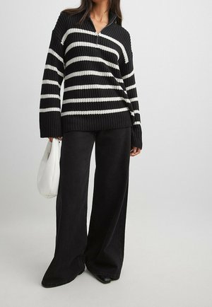 Personne portant un pull en maille rayé noir et blanc, un pantalon noir à jambes larges, tenant un sac à main blanc, debout devant un fond uni.