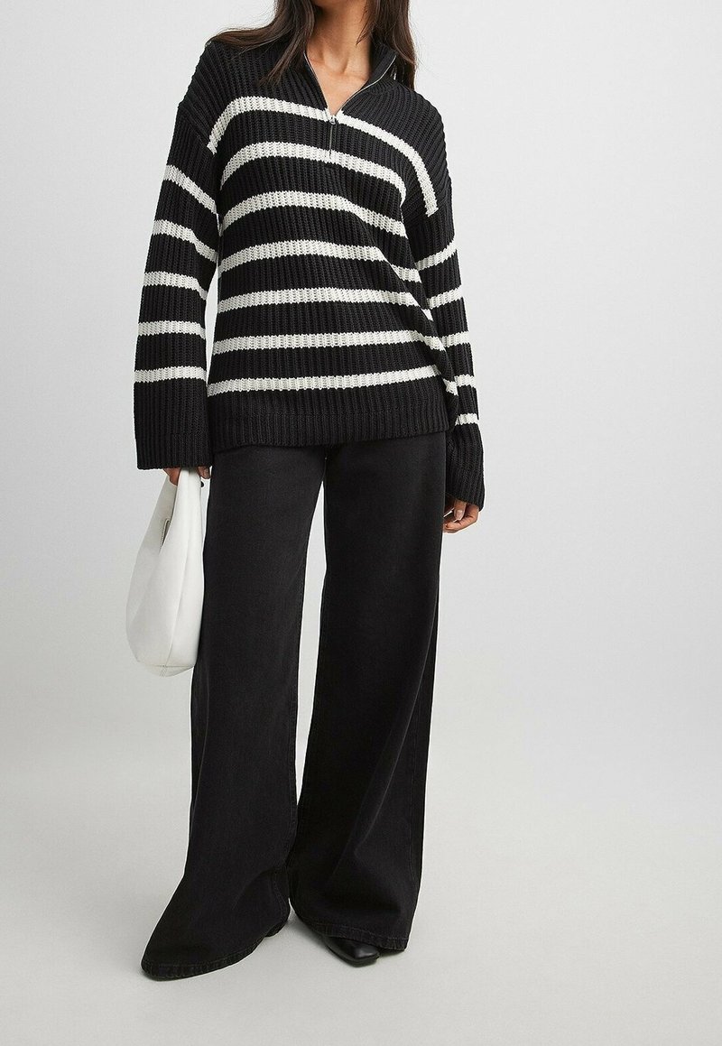 Personne portant un pull en maille rayé noir et blanc, un pantalon noir à jambes larges, tenant un sac à main blanc, debout devant un fond uni.