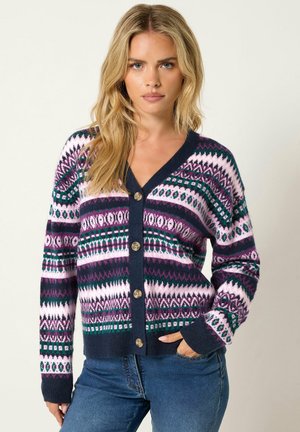 Mujer rubia que lleva un cárdigan abotonado y estampado en azul marino, rosa, verde y morado, combinado con vaqueros azules, de pie con una mano en el bolsillo.
