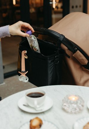 Una borsa nera su un passeggino contiene una pochette con un tappo viola. Un ciuccio rosa è attaccato alla borsa. Nei paraggi sono visibili una tazza di caffè e un pasticcino.