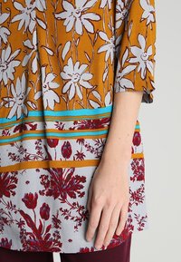 Tunique à motif floral avec un fond orange-brun, des sections gris clair et des rayures horizontales contrastantes en bleu et rouge. Texture de tissu douce.