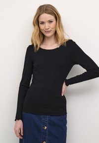 Top lungo a maniche ribbed nera con scollatura arrotondata, caratterizzato da bordi sfrangiati e una vestibilità aderente. Indossato con una gonna di denim.