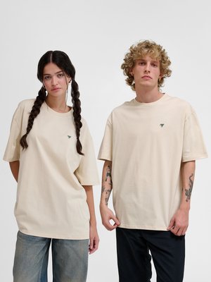 Młoda kobieta z warkoczami i młody mężczyzna z kręconymi włosami noszą beżowe, oversize t-shirty, stojąc obok siebie na jednolitym tle.