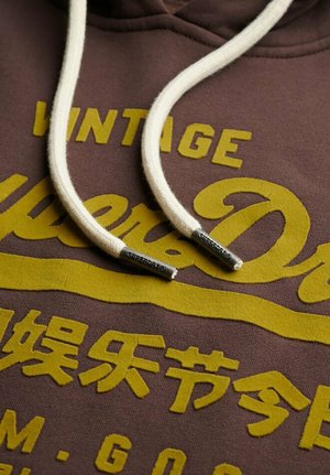 Sudadera con capucha marrón con texto amarillo "Vintage Superdry", cordones blancos con puntas metálicas etiquetadas "Superdry" y caracteres japoneses amarillos adicionales.