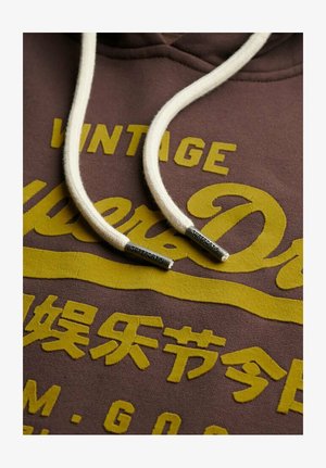 Sweat à capuche marron avec texte jaune « Vintage Superdry », cordons blancs avec embouts métalliques marqués « Superdry » et caractères japonais jaunes supplémentaires.