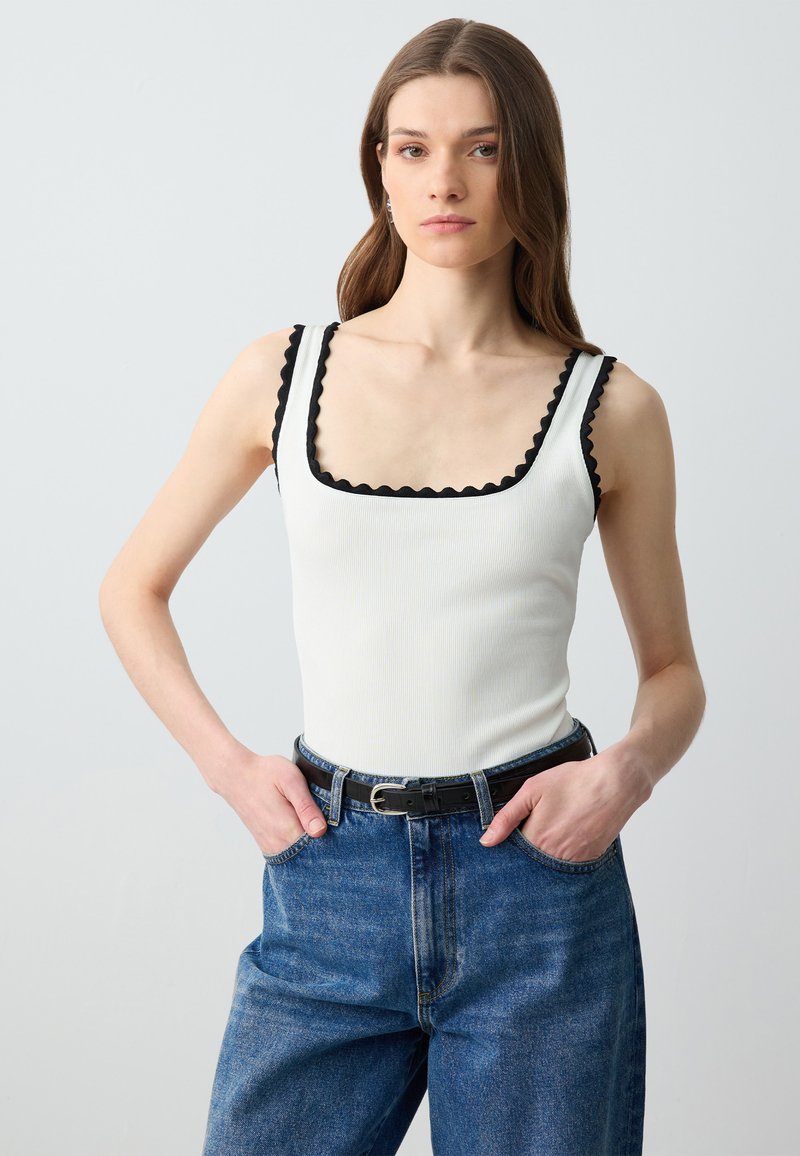 Top a canotta bianca a coste con bordi neri a scallop, abbinato a jeans blu in denim a vita alta e una cintura nera. Texture liscia, design aderente.