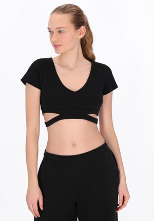 Młoda kobieta o jasnych włosach w czarnym krótkim topie z wycięciami i czarnych spodniach, stojąca na tle białego tła, patrząca w lewo.