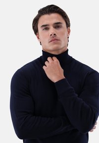 Junger Mann mit braunen Haaren, der einen dunkelblauen Rollkragenpullover trägt, mit einer Hand den Kragen hält, ernstes Gesichtsausdruck, neutraler Hintergrund.