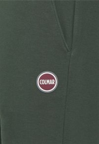 Mörkgrön sweatshirttyg med slät textur, med en rund logotyp som är märkt "COLMAR" i vitt och vinröda färger.