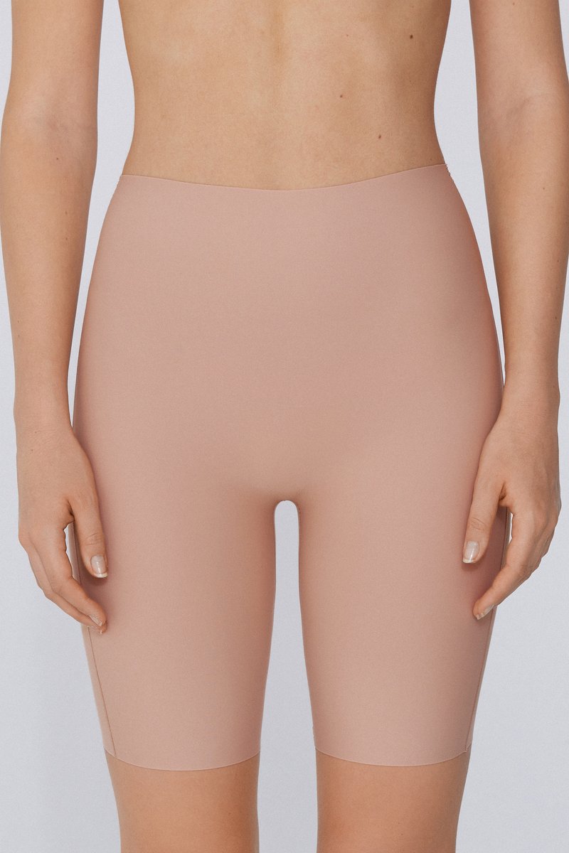 Femme portant un short gainant sans coutures, couleur beige nude, taille haute, longueur mi-cuisse, sur un fond uni.