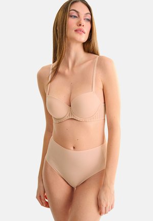 Femme aux longs cheveux portant un soutien-gorge couleur chair à fines bretelles et une culotte taille haute assortie, debout sur un fond clair uni.