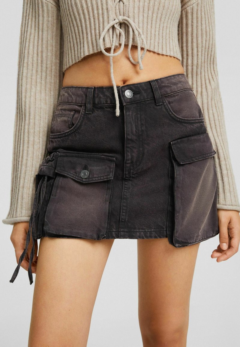 Femme portant une mini-jupe en jean noire avec de grandes poches avant et une fermeture à boutons, associée à un pull court côtelé beige.