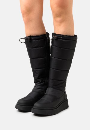 Bottes noires matelassées montant jusqu'au genou, avec semelles épaisses et fermetures à cordon réglable en haut, portées sur des jambes nues.