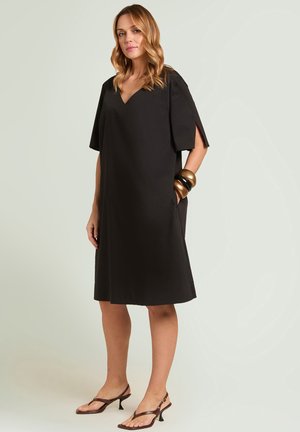 Femme portant une robe noire ample jusqu'aux genoux avec des manches fendues aux coudes, des sandales marron à talons et des bracelets imposants à un poignet.
