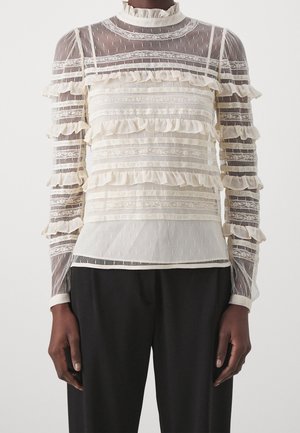 Sie trägt eine durchsichtige cremefarbene Spitzenbluse mit horizontalen Rüschen und hohem Kragen, kombiniert mit lockeren schwarzen Hosen, vor einem schlichten Hintergrund.