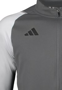 Graue Sportjacke mit hohem Kragen, schwarzen Akzenten und Reißverschluss. Mit schwarzem Adidas-Logo auf der linken Brust und kontrastierendem Schulterdesign.