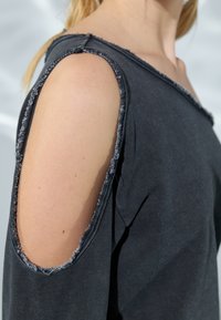 Chemise noire avec découpes aux épaules dénudées bordées de franges, en tissu doux. L'encolure est large et légèrement décalée sur les épaules.