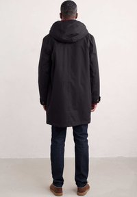 Veste extérieure noire avec une grande capuche, de longueur longue et des poignets boutonnés, associée à un jean bleu foncé et des chaussures marron.