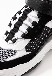 Černobílé tenisky s síťovaným svrškem a perforovanými detaily, silnou podrážkou, bezšňůrovým provedením a popruhem s embosovaným logem.