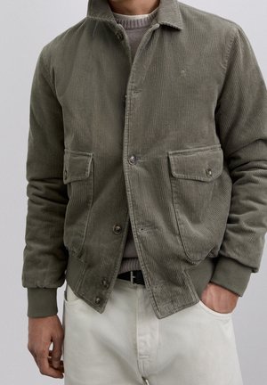 Uomo che indossa una giacca di velluto a coste verde oliva con tasche con bottoni sopra a un maglione grigio e pantaloni bianchi, con una mano in tasca.