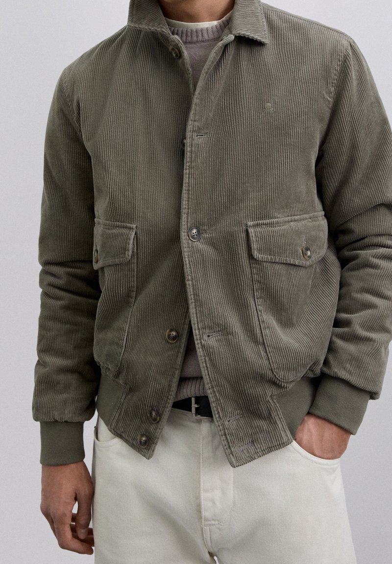 Homme portant une veste en velours côtelé vert olive avec des poches boutonnées sur un pull gris et un pantalon blanc, une main dans la poche.