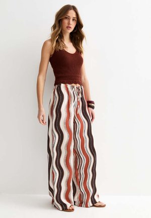 Vrouw met los golvend haar draagt een mouwloze bruine gebreide top en wijde broek met verticale golvende strepen in bruin, roestkleur en wit.