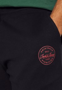 Black sweatpants featuring a textured fabric, elastic waistband, and a red circular logo with "Jack & Jones" and "Established 1990."