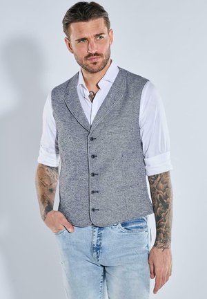 Mann mit hochgekrempelten weißen Hemdsärmeln, grauer Knopfweste, blauer Jeans und tätowierten Armen, der mit einer Hand in der Tasche steht.