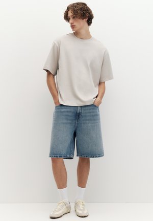 Ung mand med krøllet hår iført en løs, lys beige t-shirt, blå denimshorts, hvide sokker og cremefarvede sneakers, der står op ad en hvid væg.