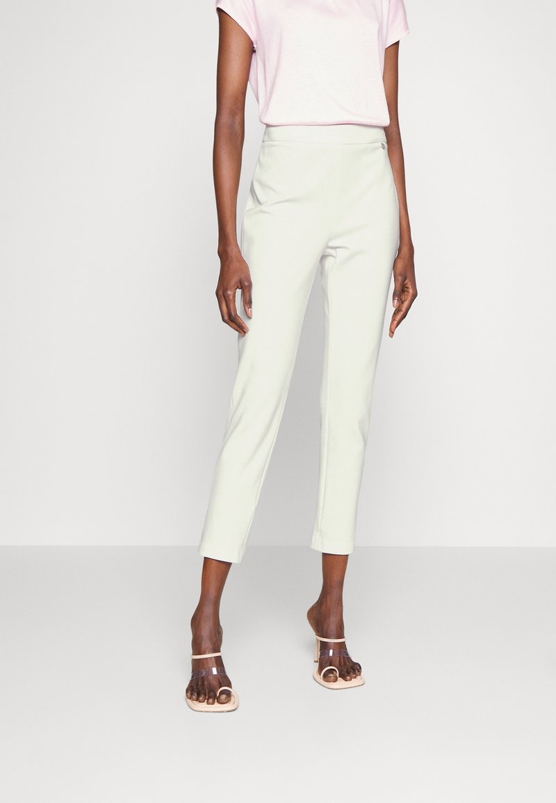 Personne portant un haut rose clair à manches courtes, un pantalon blanc court taille haute et des sandales à talons à brides transparentes sur fond uni.